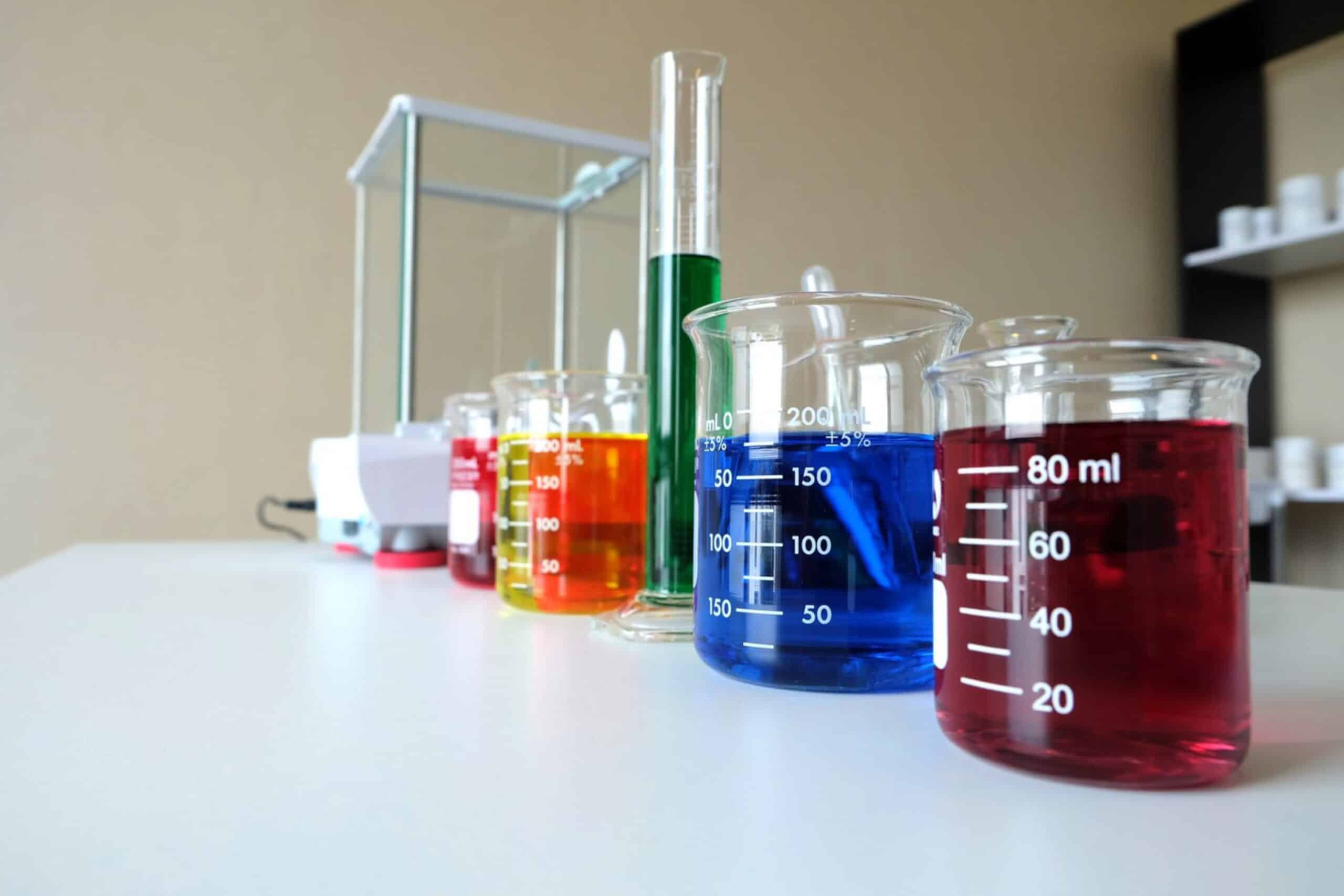 A laboratory countertop displays colorful beakers of liquids: red, blue, yellow, green, and orange, alongside a glass scale and equipment.
