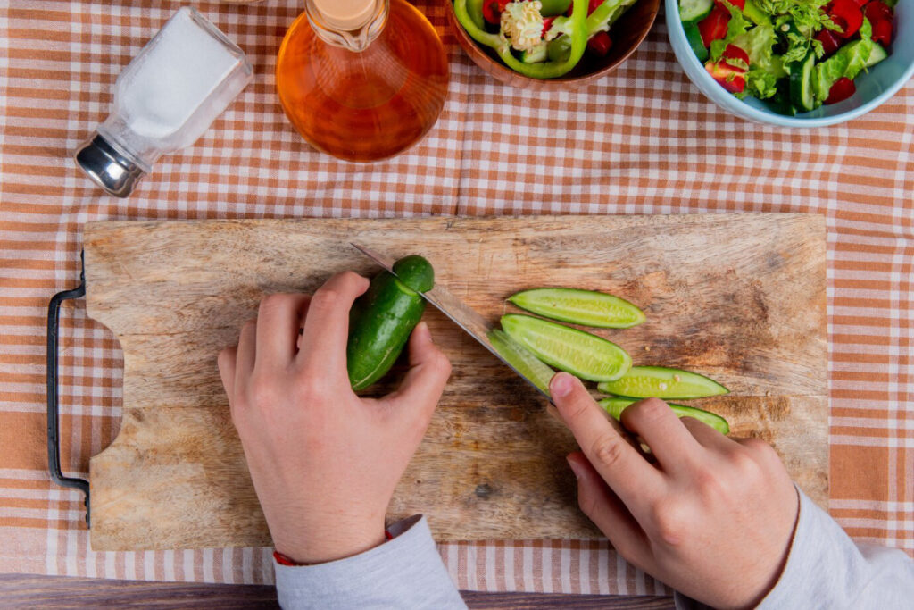 A person slices a cucumber on a wooden cutting board with a knife, surrounded by salad ingredients, salt, and oil on a checkered cloth.