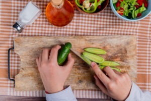 A person slices a cucumber on a wooden cutting board with a knife, surrounded by salad ingredients, salt, and oil on a checkered cloth.