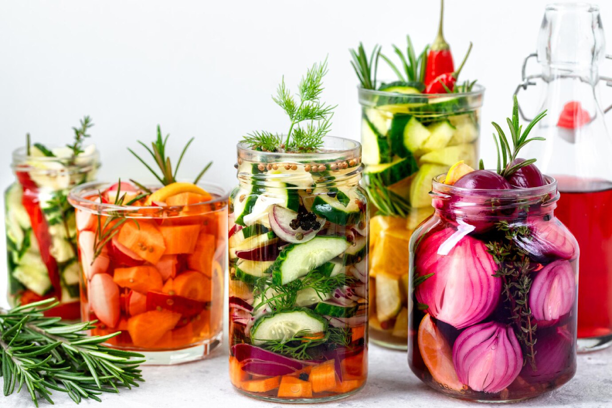 A variety of colorful pickled vegetables in glass jars, featuring cucumbers, carrots, and red onions, garnished with herbs.