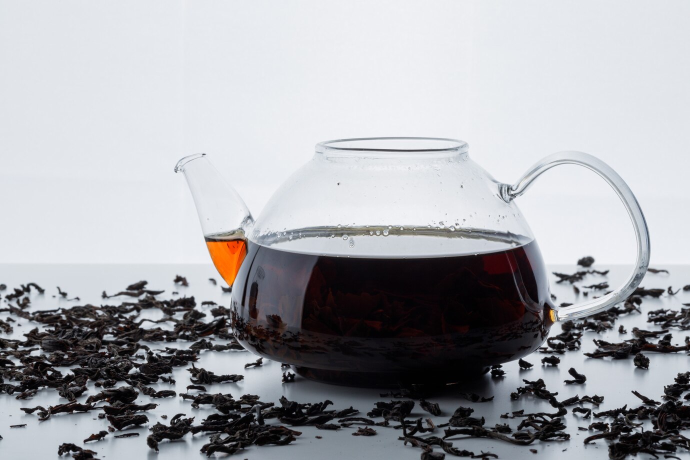 A glass teapot filled with dark brown tea sits on a white surface surrounded by loose black tea leaves.