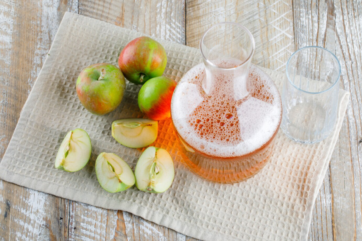 A glass decanter filled with bubbly apple juice, surrounded by fresh apples and sliced apples on a textured cloth.