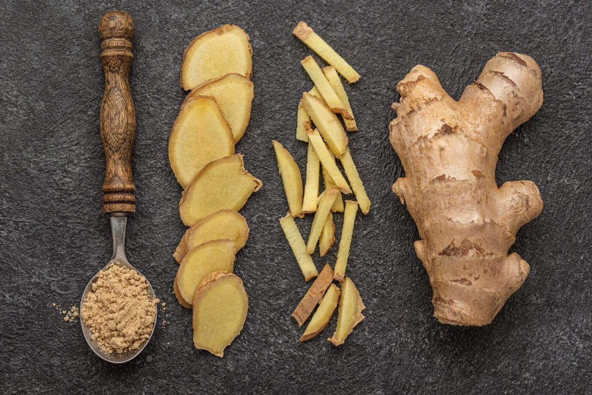 A spoonful of ground ginger beside slices, thin strips, and a whole ginger root on a dark textured background.