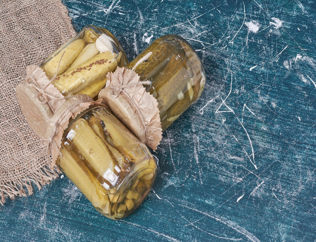 Three jars of pickles wrapped with brown paper lids, resting on a textured blue surface next to a piece of burlap.