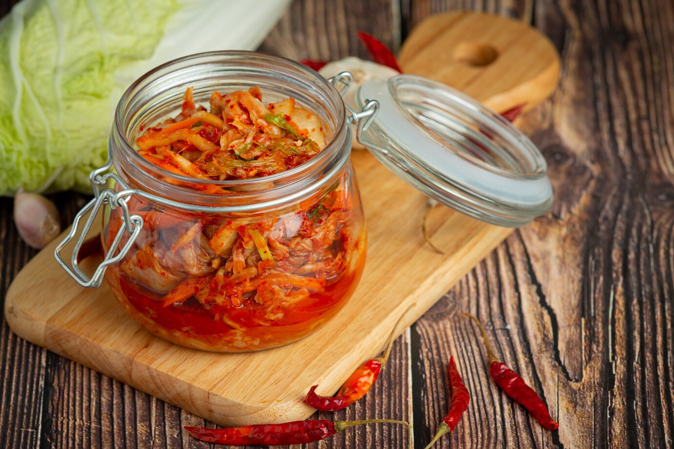 A glass jar of vibrant kimchi sits on a wooden cutting board, surrounded by dried red chilies and a cabbage in the background.