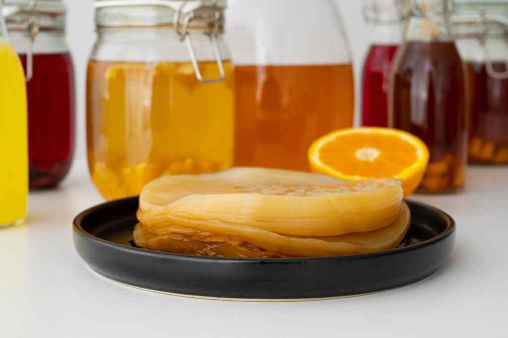 A black plate holds a kombucha SCOBY, with jars of colorful brewed teas and an orange slice in the background.